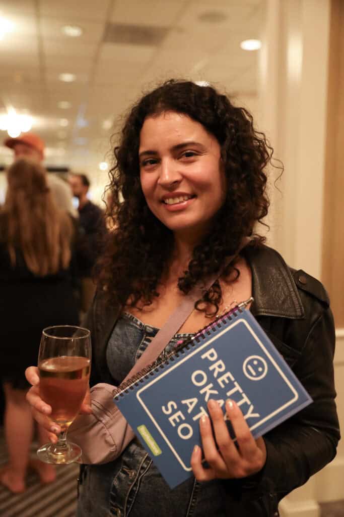 A woman holding a glass in one hand and a Pretty Okay At SEO notebook in the other hand.